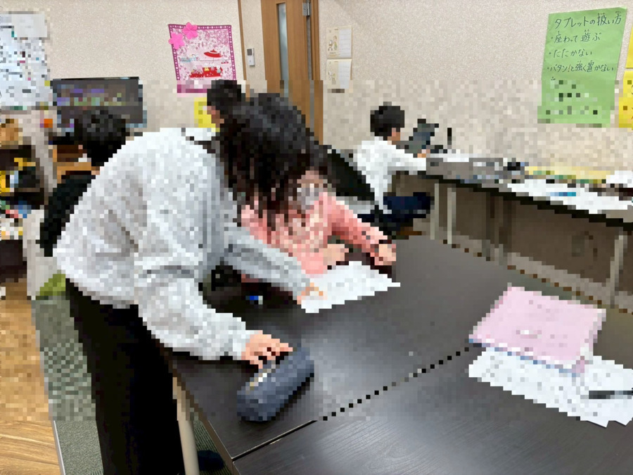 こどもデイサービス「アレ・オリヴィエ」（児童指導員/指導員の求人）の写真3枚目：アレ・オリヴィエでスタッフと学習している利用者。その奥でタブレットをする利用者。
